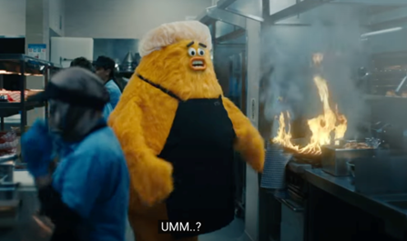 A yellow, costumed mascot working in a fast-paced kitchen is being pressured to hurry and finish a customer's order. Becoming overwhelmed, the mascot catches a kitchen towel on fire and utters a panicked "Umm...? as an expression uncertainty about what to do next.